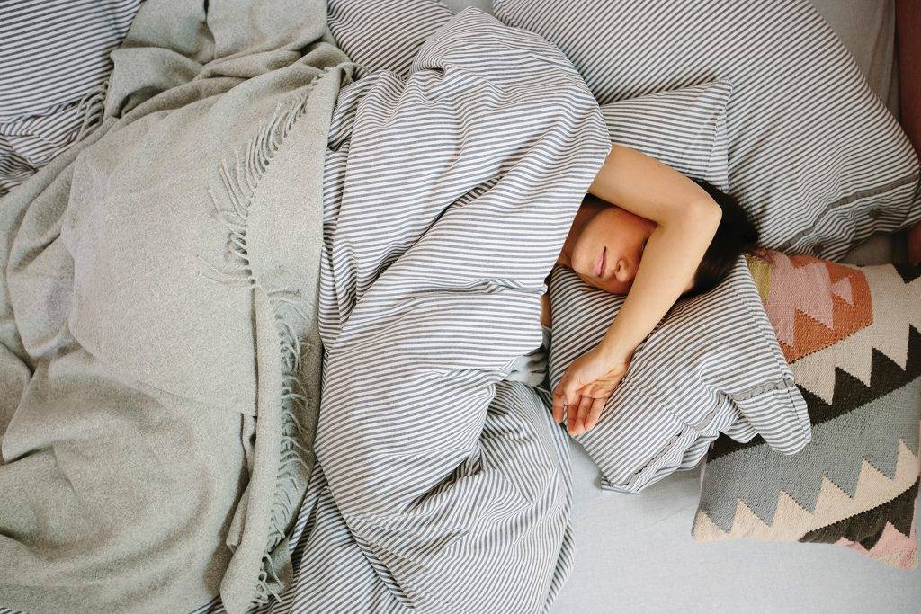 From above young female covering eyes with arm while lying on soft bed under warm blanket and sleeping in morning at home